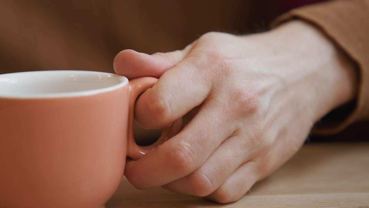 Primo piano di una mano che si allunga per afferrare una tazza di ceramica rosa vuota su un tavolo di legno. La scena presenta un'illuminazione soffusa con lo sfondo sfocato per enfatizzare l'interazione tra le dita del soggetto umano