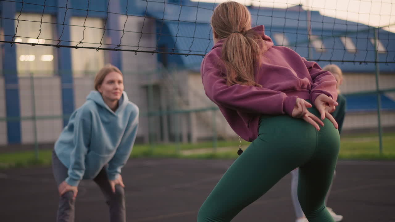 vista trasera de una dama ligeramente inclinada haciendo gestos, con otras dos damas borrosas en el fondo en postura similar durante una actividad de entrenamiento físico al aire libre, participando en trabajo en equipo