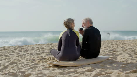 long shot of a senior couple in wetsuit drinking tea on beach after surf training