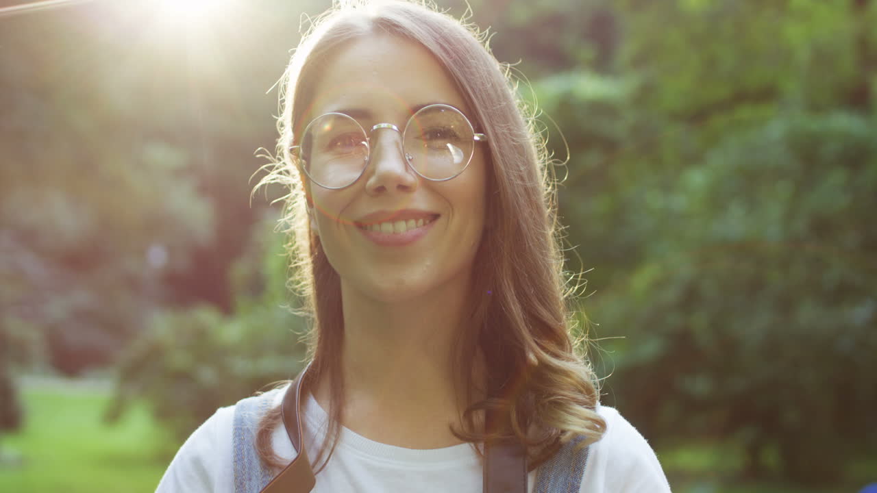 Close-up view of a beautiful young woman in glasses taking photos in the park with a old vintage photo camera and smiling at camera