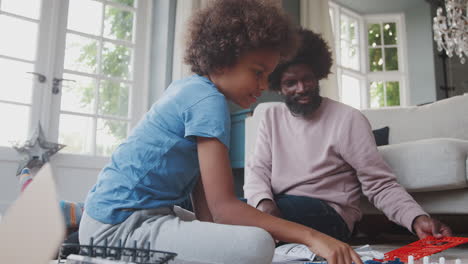 middle aged black father sitting on the floor at home helping his pre teen son with the parts of a toy construction kit, close up, low angle