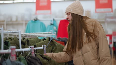 a young girl wearing a brown beanie and matching jacket browses through a clothing rack in a mall, focusing on a green jacket. the background features a variety of colorful clothing