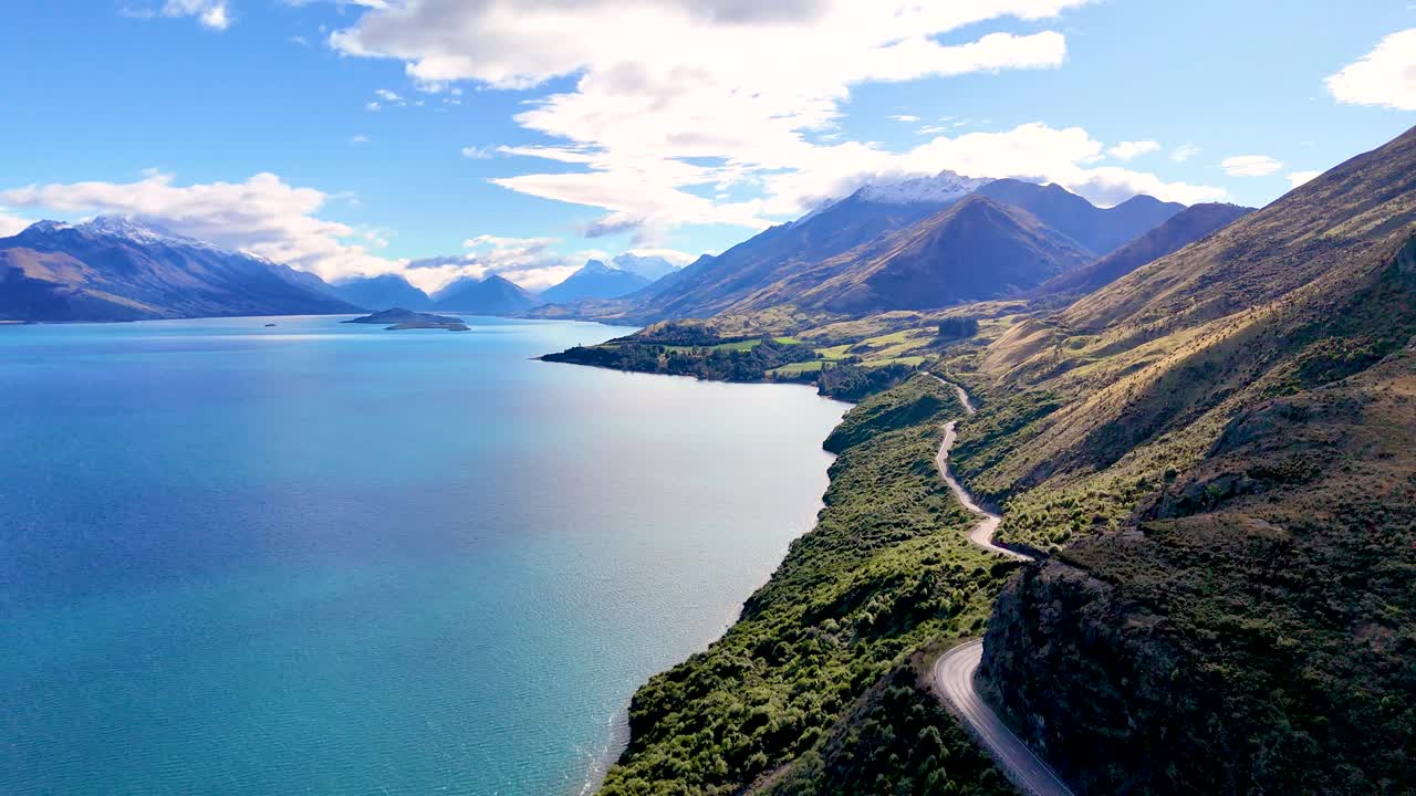 Drone glides above winding lakeside road, revealing mountains, blue water, and lush greenery