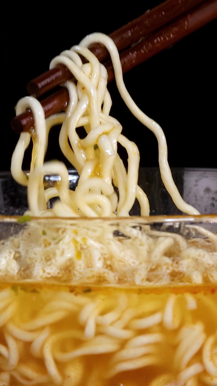 Bacchette afferrano e sollevano noodles istantanei cotti da una ciotola di vetro fumante di brodo, con inquadratura ravvicinata, illuminazione drammatica e uno sfondo nero.