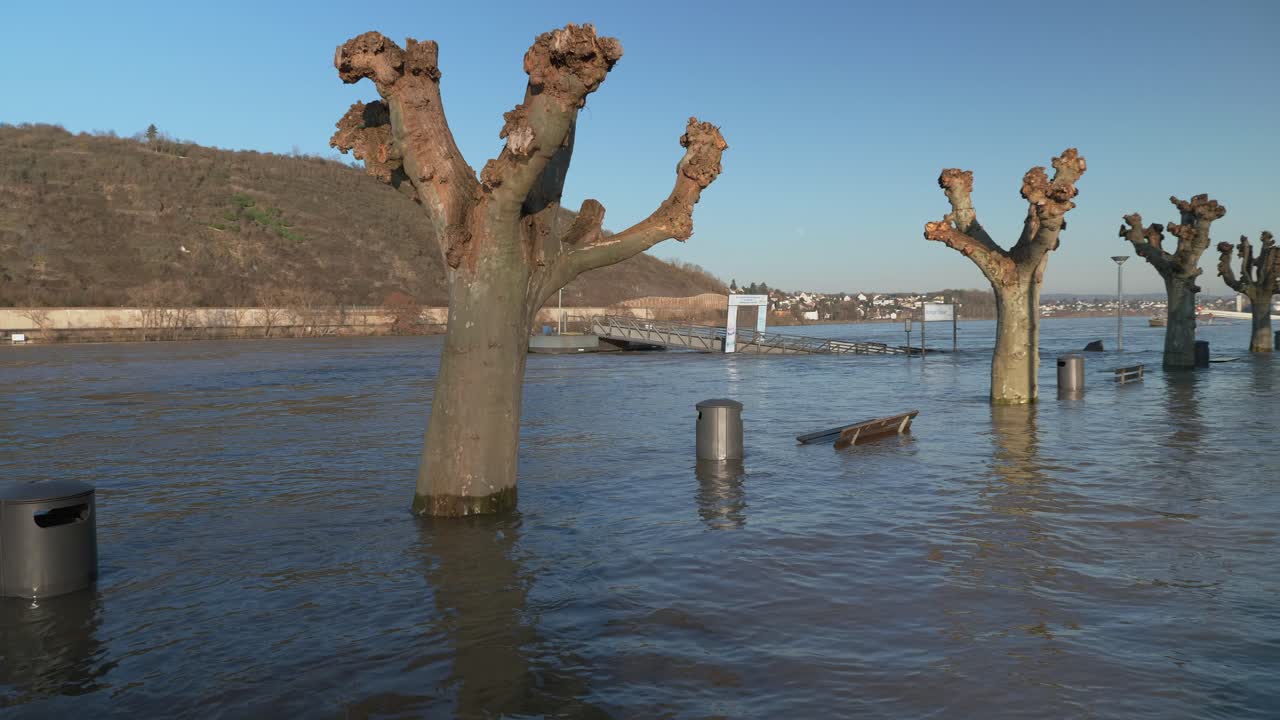 Benches, trash cans, street lamps and trees standing in the flood water with river and city in the background. Wide angle shot, panning from right to left.