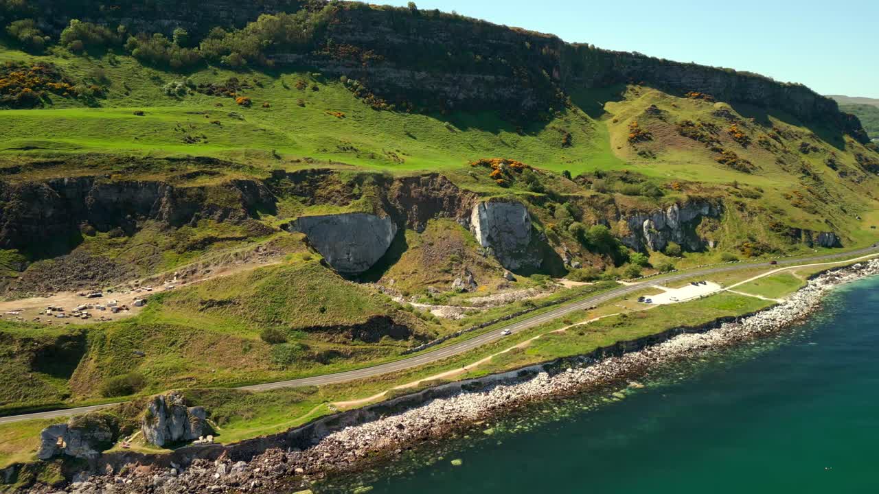 Aerial video of the Causeway Coastal Route in County Antrim, Northern Ireland, UK, on a bright and sunny day. Cars pass below the green Irish cliffs. Filmed in 4K, 60FPS and with Rec709 color.