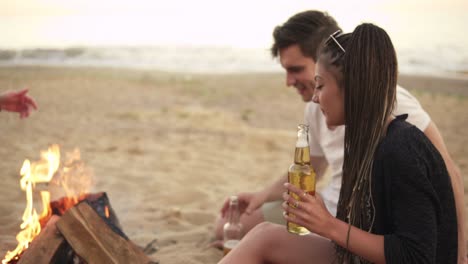 joven abre botellas, bebe cerveza y con amigos sentados junto al fuego en la playa. amigos divirtiéndose. filmado en 4k.