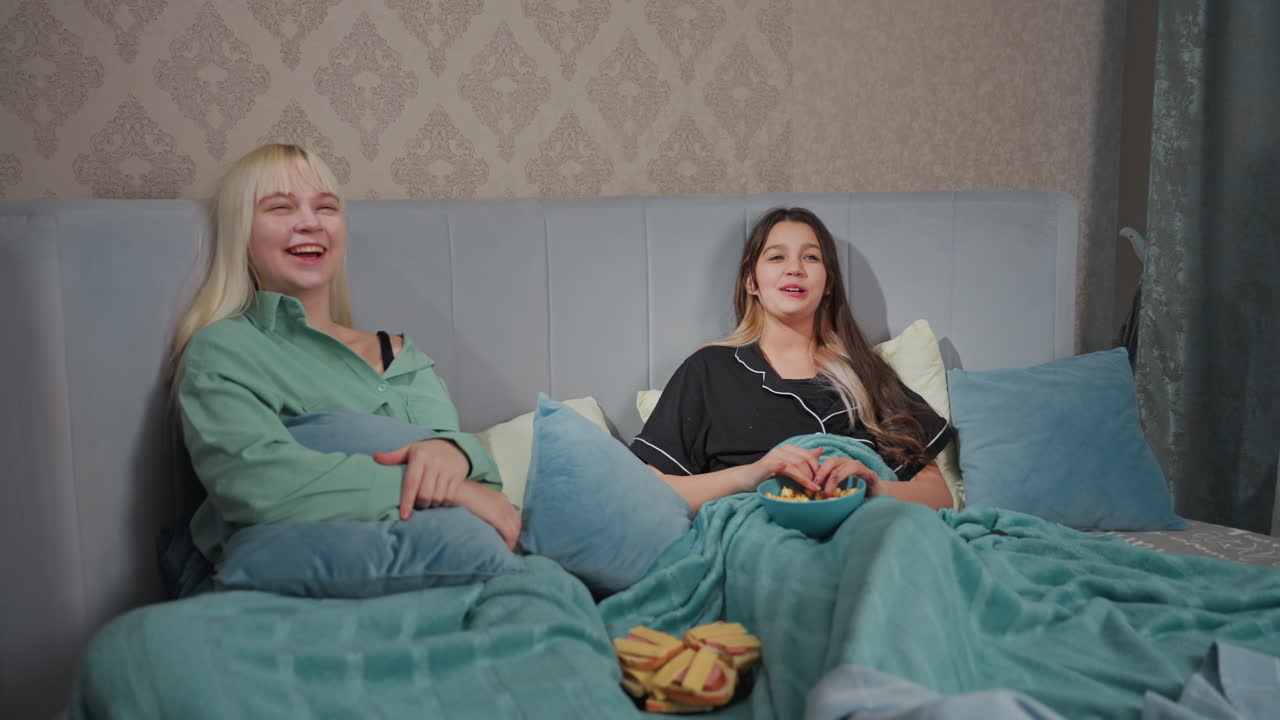 Two girls lie in bed under teal blanket, laughing and enjoying snacks in relaxed setting, one holds bowl of popcorn, other lounges with pillow