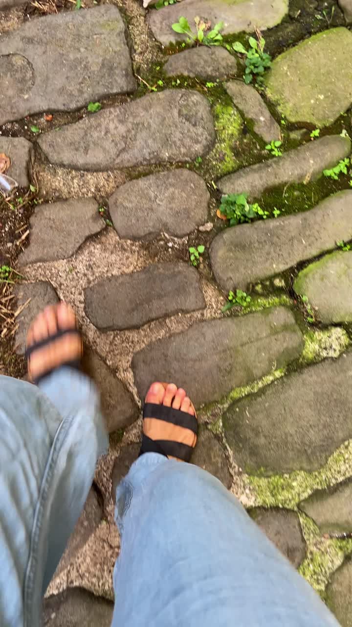 Female traveler or hiker tourist enjoying nice walk on old ancient stone road with green vegetation. Walking with sandals and jeans in historic town square in Europe. Medieval rock path. Exploring