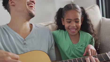 Glücklicher-Gemischtrassiger-Vater-Und-Tochter,-Die-Auf-Dem-Sofa-Sitzen-Und-Gitarre-Spielen