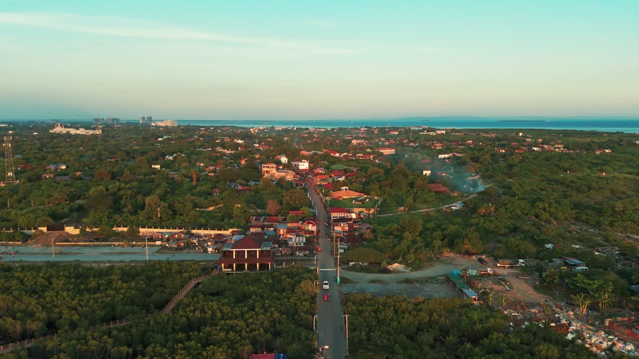Aerial drone shot of a rural road lined with houses and trees, leading into a dense Philippine township and coastal skyline during golden hour. Ideal for city growth or transport themes