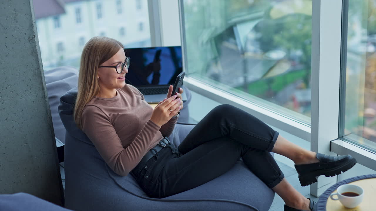 Relaxed long-haired lady wearing glasses sits in bean bag chair. Woman looking at her phone and then taking selfie. Blurred cityscape at backdrop.