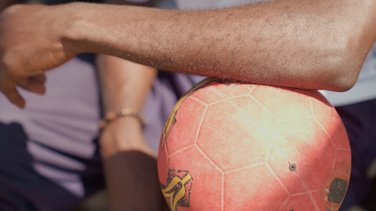 Close-up de braço negro sobre bola de futebol, com gesto de apontar e colegas de equipe no banco, em conversa casual pré-jogo, luz solar enfatizando a superfície de borracha texturizada, energia e antecipação de equipe urbana.