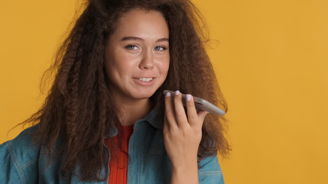 una mujer caucásica de cabello rizado grabando un mensaje de voz en su teléfono inteligente.