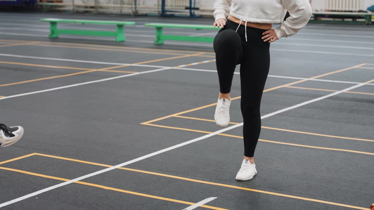 Riscaldamento del gioco di gambe su campo da badminton: atleta donna bianca che pratica sollevamenti precisi delle ginocchia e equilibrio su un campo indoor segnato con coni a scaletta e allenatore nelle vicinanze, con intensità concentrata e ritmo costante.