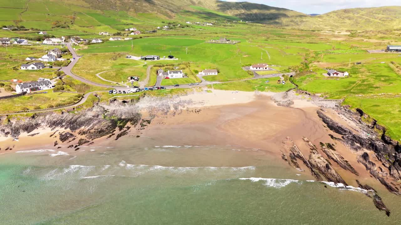 Aerial shot begins over a surfer at sea, rising to reveal Ireland’s rugged coast, scattered homes, and lush green hills in the distance.