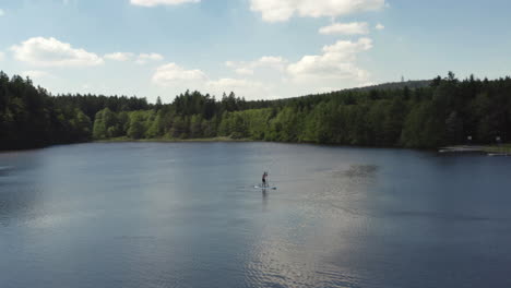 a lone person paddling a stand up paddleboard across a calm pond on vacation, aerial orbit