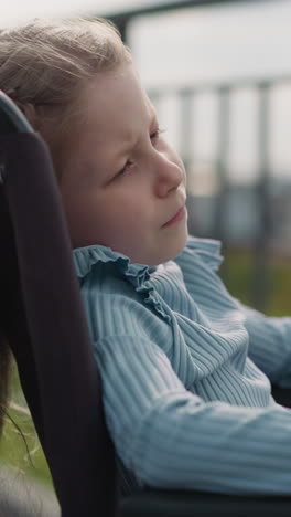 preschooler girl with paralysis sits in wheelchair looking at city on blurred background. tranquil girl with disability enjoys sunny spring weather closeup