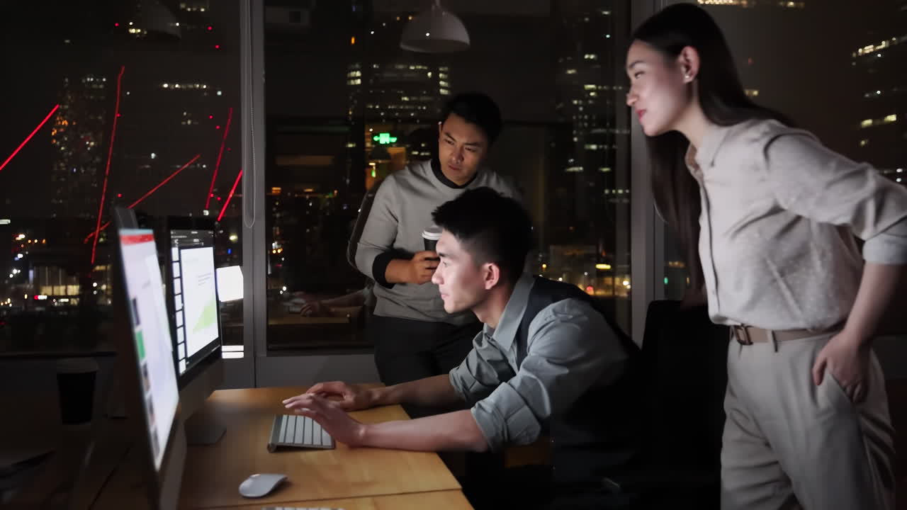 Three male and female business colleague office workers in front of computers late at night creative session, exchanging ideas and solution to meet a project deadline, with city lights in background