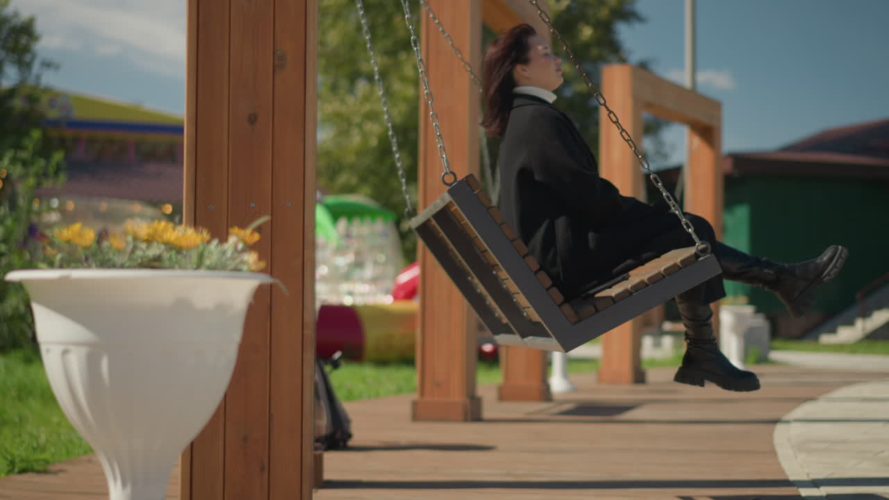 스타일리시한 검은 코트와 부츠를 입은 여성은 빛이 비치는 공원에서 녹색과 장식 꽃으로 둘러싸인 나무 스에 휴식을 취하고, 밝은 하늘 아래 나무 구조물을 배경으로합니다.