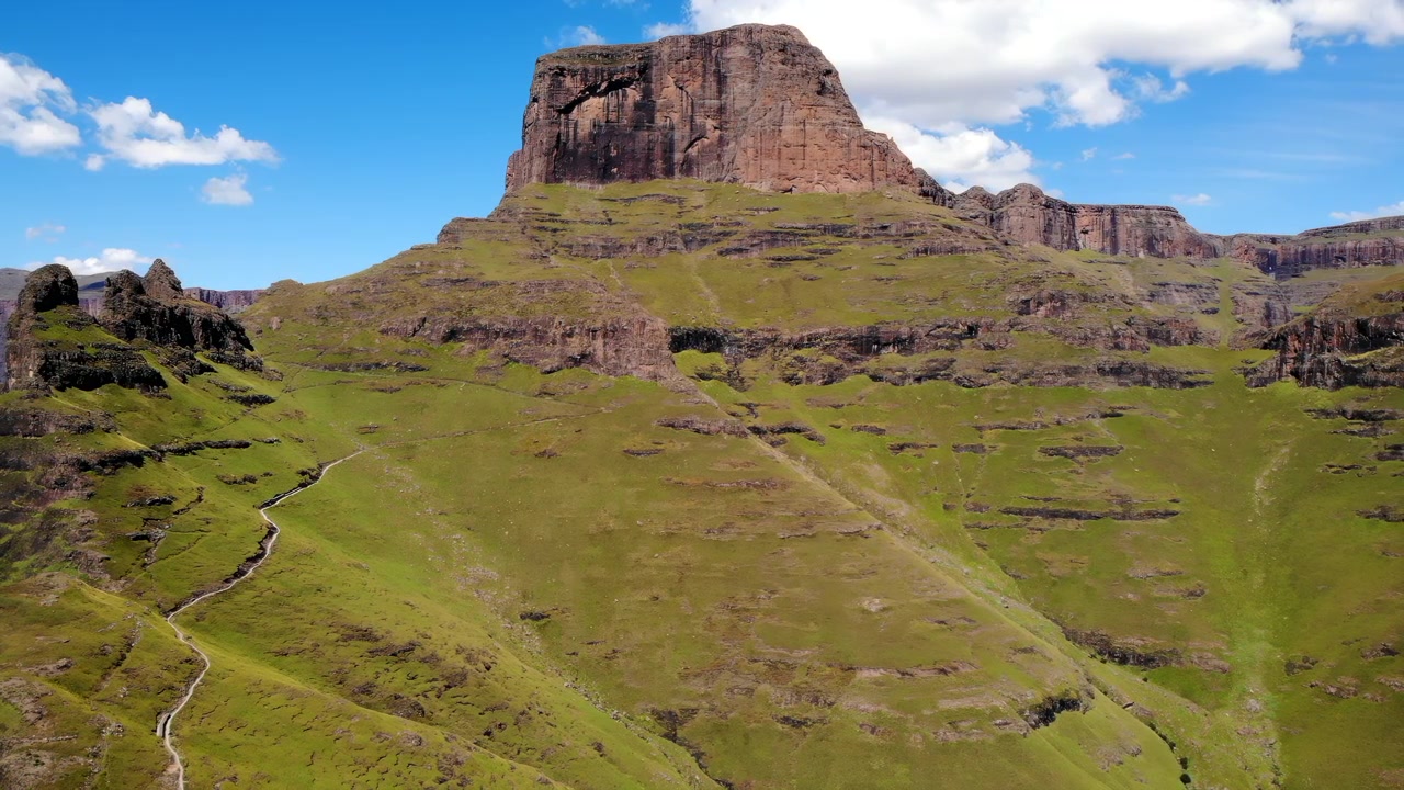 Ampia vista panoramica di una montagna aspra che si erge su pendii erbosi e sentieri tortuosi, con luce diurna intensa, nuvole sparse e una natura selvaggia aperta che si estende.