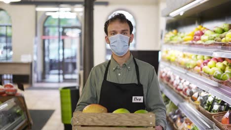 Portrait-of-handsome-man-in-face-mask-and-apron-going-ahead-in-food-store-with-box-of-fruits-indoors