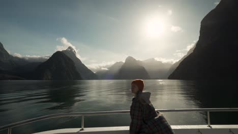 Girl-on-a-boat-cruise-taking-a-scenic-picture-of-sun-beaming-through-steep-mountain-faces-and-islands-in-New-Zealand's-Milford-Sound