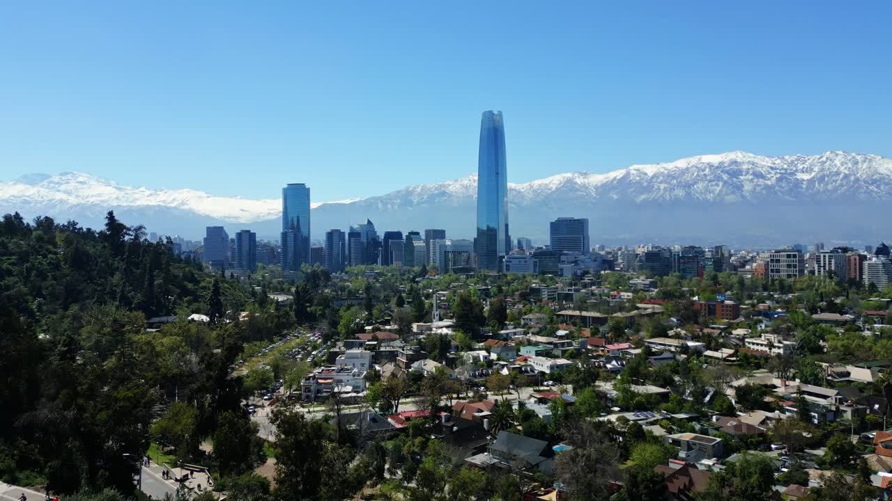 Drone aerial forward approach over treelined hills and rooftops toward the Costanera skyscraper, with snowcapped Andes on the horizon in clear daytime light. Santiago, Chile