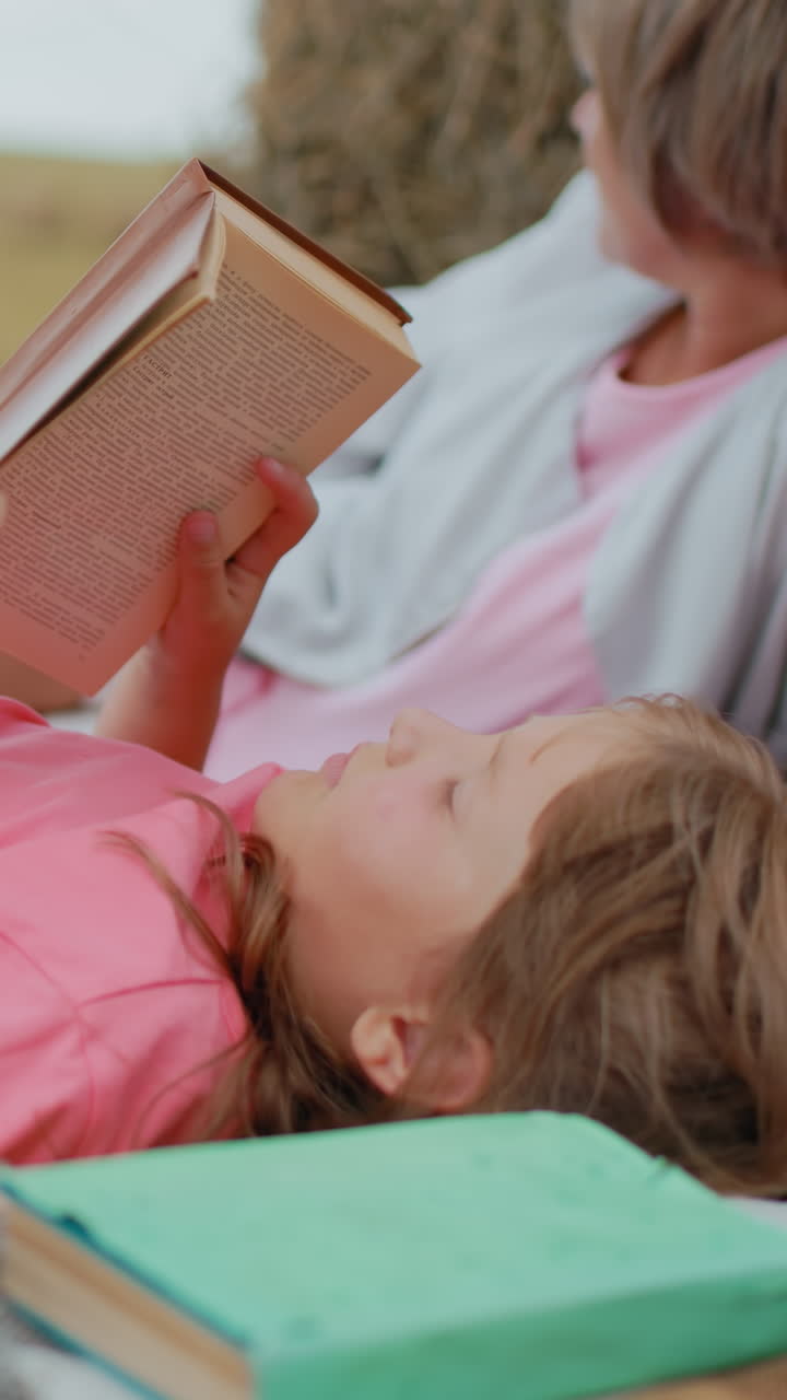 primer plano de una niña con camisa rosa leyendo su libro mientras está acostada en una manta de picnic, libro verde cerca de la cabeza, familia relajándose con el perro en la correa y disfrutando de un tiempo tranquilo al aire libre en el campo