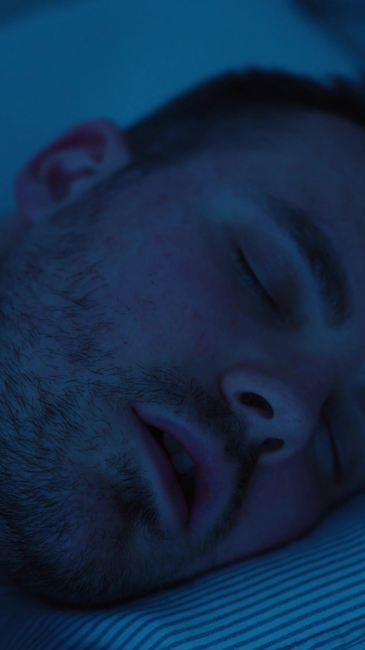 Video verticale ravvicinato di un uomo che dorme a bocca aperta sotto una luce notturna blu, evidenziando la respirazione orale, il russare, problemi di sonno, riposo, affaticamento, l'ora di coricarsi e preoccupazioni per la salute notturna.