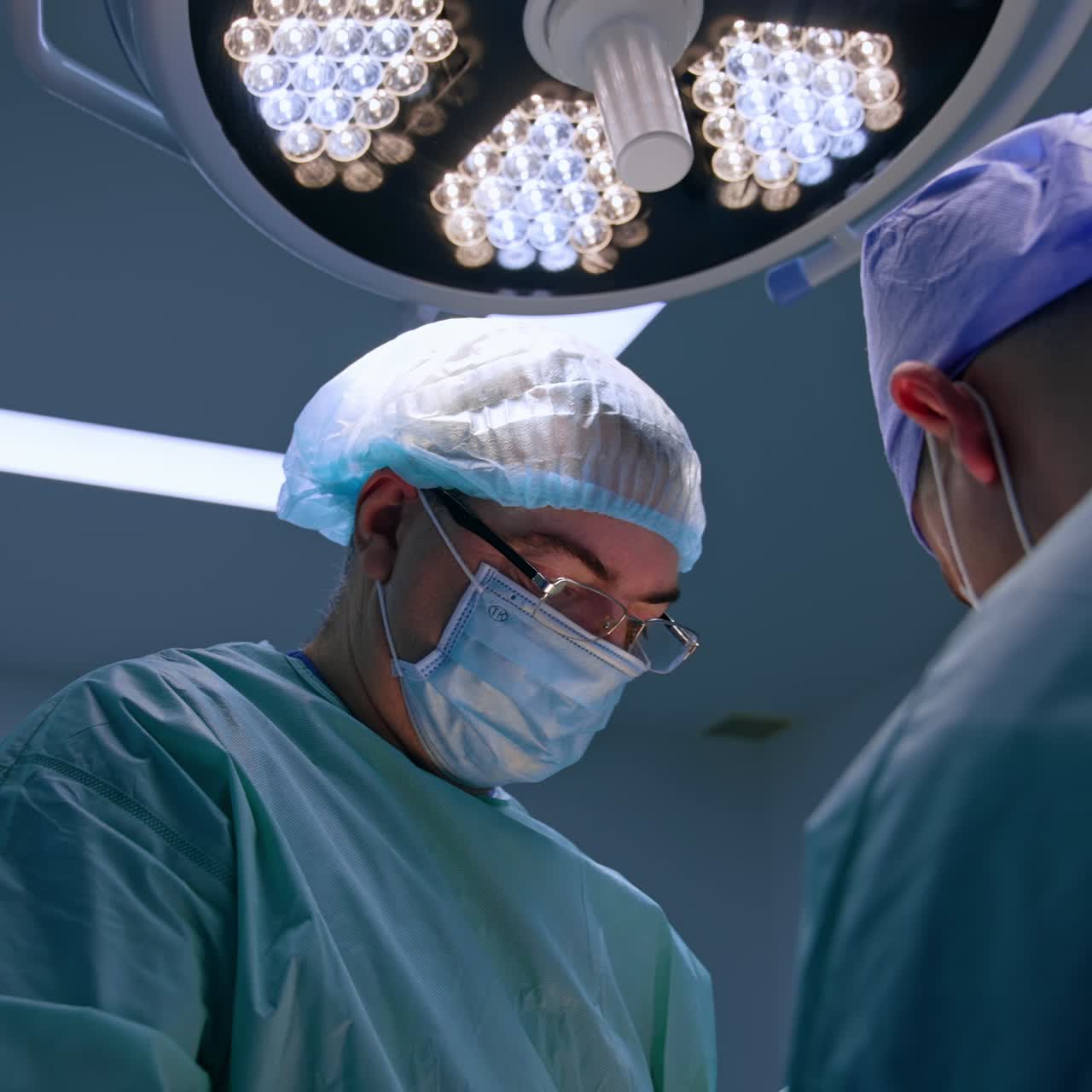 Skilled male surgeon in eyeglasses and mask works at operation. Portrait of a doctor working in tam and focused on procedure. Low angle view