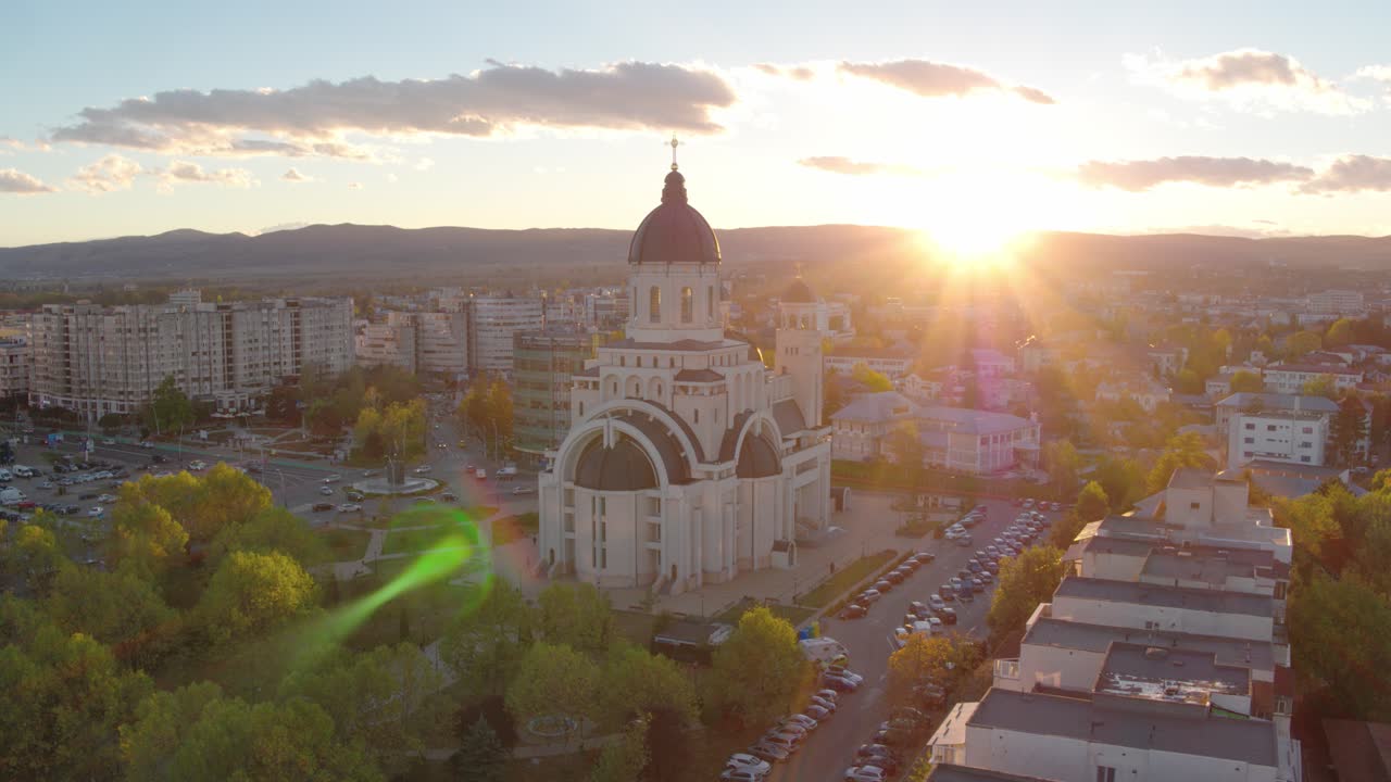 A slow-panning aerial shot of an Orthodox cathedral in Bacau, Romania. The setting sun creates a spectacular lens flare over the autumn park and cityscape