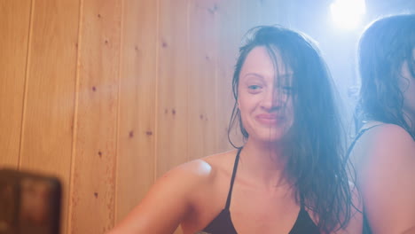 close up of woman recording video with smartphone against warm wooden panel backdrop, soft backlight illuminating wet hair and smile, candid beauty content creation