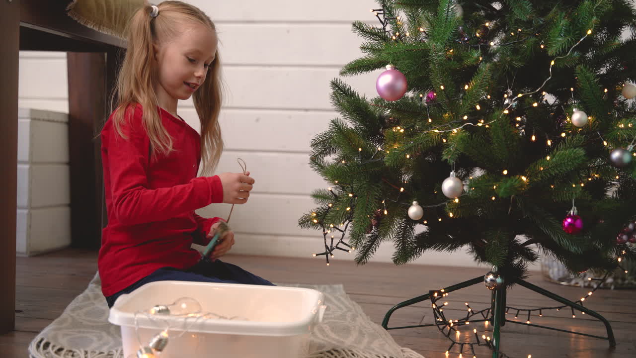 小金发女孩跪在地板上从盒子里拿起圣诞装饰并挂在圣诞树上