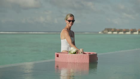 Woman-in-white-swimsuit-drinking-fruit-smoothie-from-floating-breakfast-while-sitting-on-infinity-pool-edge