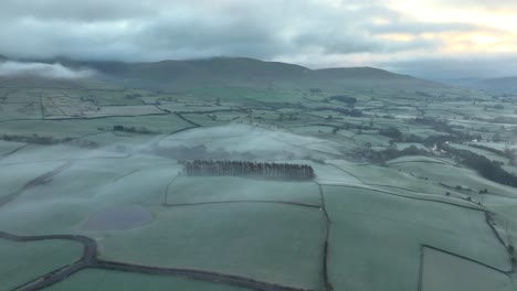 Patchwork-green-fields-with-fog-and-mist-drifting-over-them-and-low-mountains-in-distance-at-dawn-in-winter