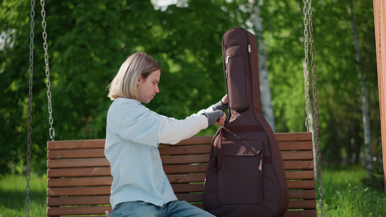 Donna caucasica bionda che apre la custodia di una chitarra su una panchina a dondolo in legno in un parco soleggiato, ispeziona lo strumento e regola la tracolla, si prepara per una sessione acustica all'aperto con espressione calma e concentrata, indossa jeans di denim