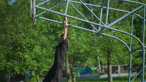 A young boy wearing a black outfit and glasses swings slowly on outdoor equipment, with lush green trees and a fence in the background