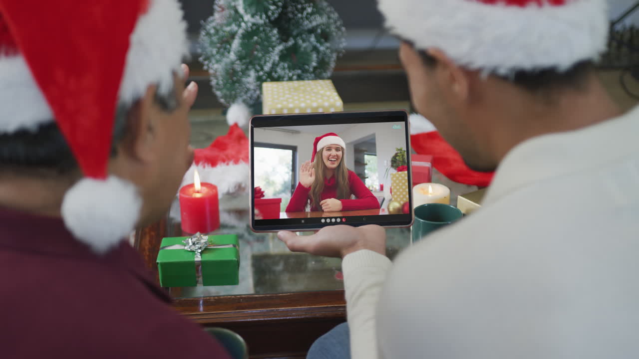 padre y hijo biraciales agitando y usando una tableta para una videollamada de navidad con una mujer en la pantalla