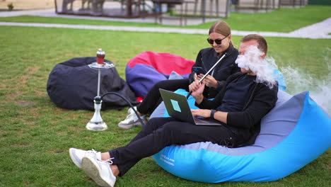young people in black clothes lie in bean bag chair relaxing outdoors. man is working on laptop and smoking hookah and woman looks at her phone.