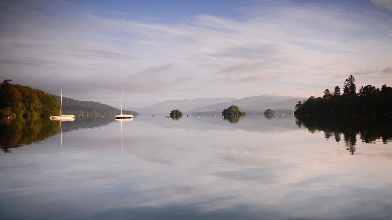 Reflection Of The Sky On The Clear Waters Of The Lake Surrounded By Green Forest And Mountains In Lake District, England. -wide shot