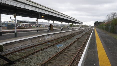 Empty-railway-platform-with-tracks,-waiting-area,-and-graffiti-wall-in-background,-under-an-overcast-sky