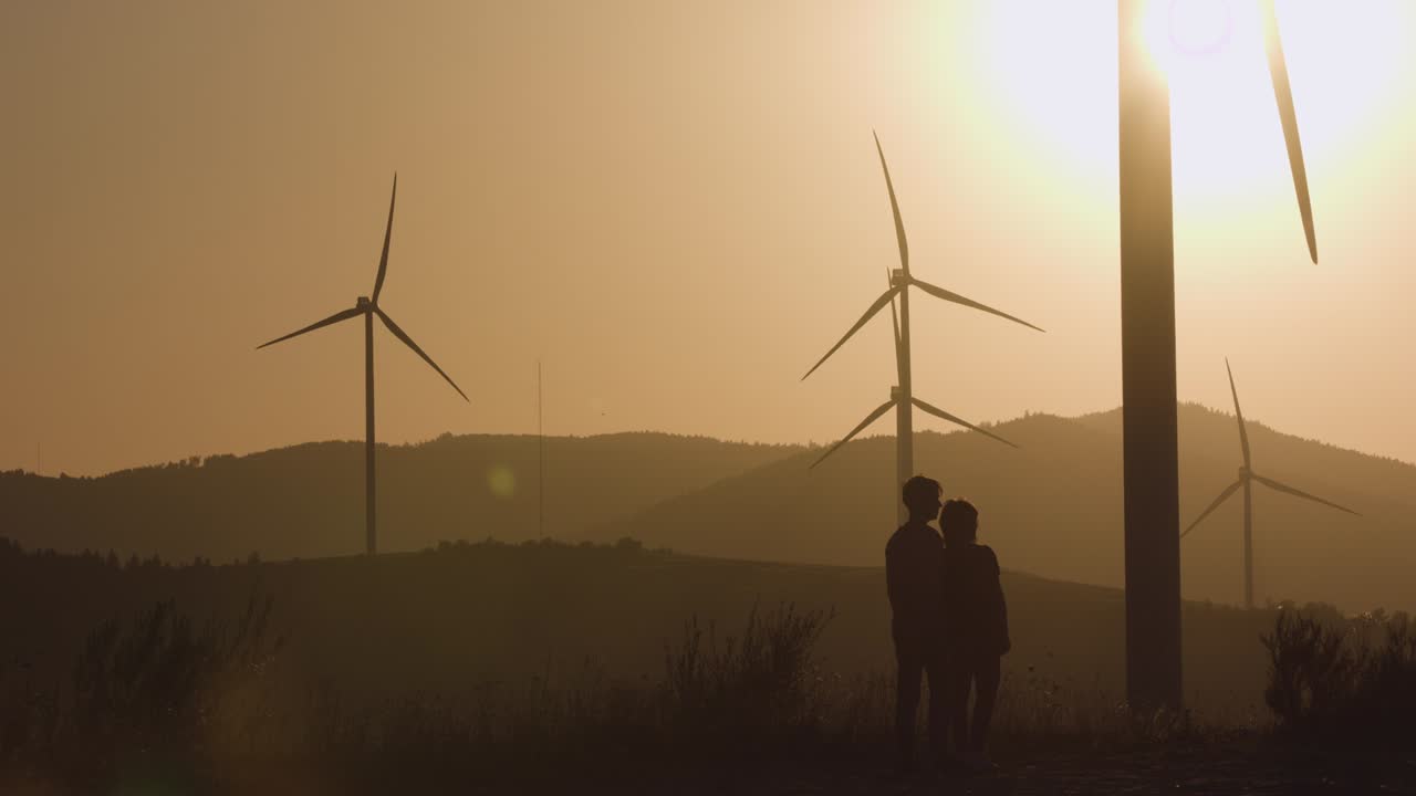 dos hombres jóvenes de pie cerca de las turbinas eólicas apuntando su mano hacia adelante. tiro de silueta. puesta de sol
