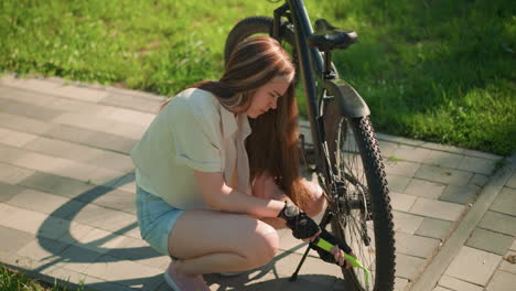 una mujer joven con guantes negros se agacha en un camino pavimentado, centrada mientras bombea aire en el neumático trasero de su bicicleta, con un telón de fondo de hierba verde y árboles