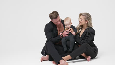 caucasian couple in black suits sit on the floor with their infant son. happy young family portrait in studio. white backdrop.