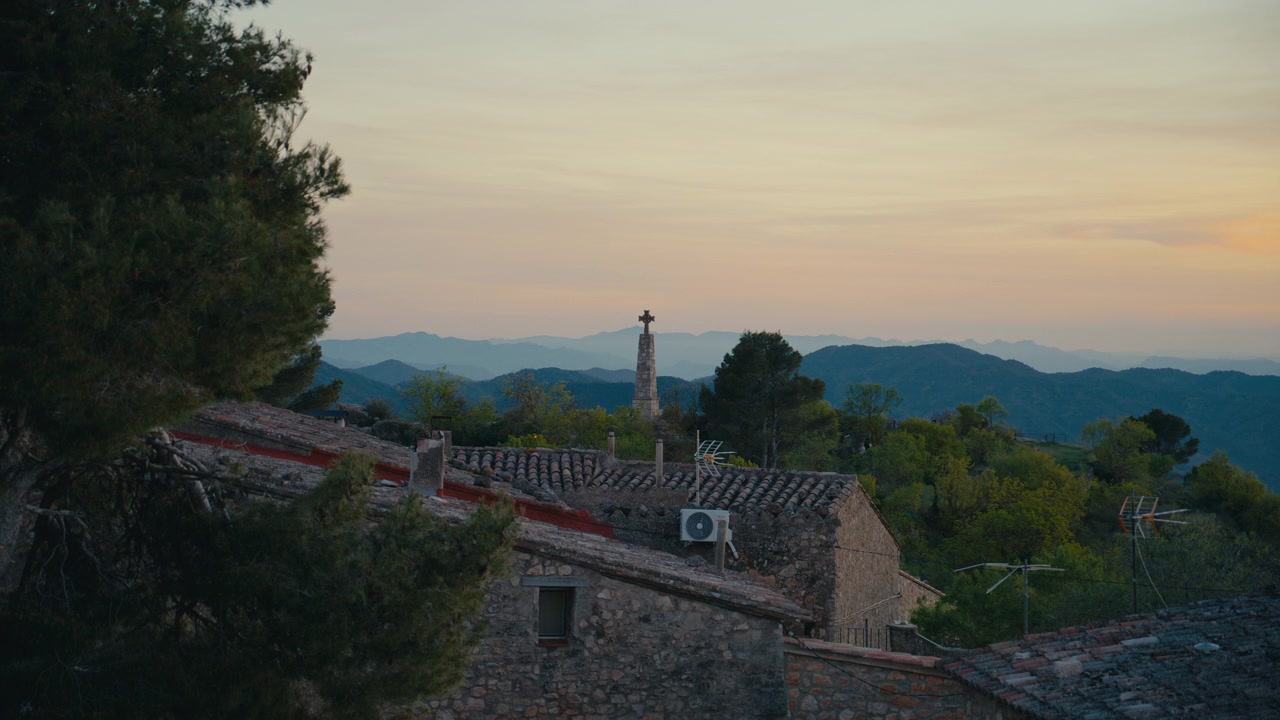 Un monumento slanciato si erge sopra i tetti di Siurana, Spagna, con catene montuose a strati e una morbida luce serale che conferiscono alla scena un senso di calma e maestosità.