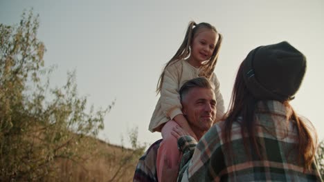 Un-Hombre-Moreno-De-Mediana-Edad-Con-Un-Poco-De-Pelo-Gris-Se-Comunica-Con-Su-Esposa-Con-Un-Sombrero-Y-Una-Camisa-A-Cuadros-Mientras-Su-Pequeña-Hija-Se-Sienta-Sobre-Sus-Hombros-Durante-Su-Picnic-Fuera-De-La-Ciudad-En-Verano.