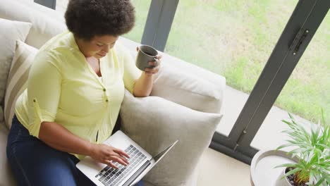 Video-of-plus-size-african-american-woman-sitting-on-sofa-with-coffee-and-laptop