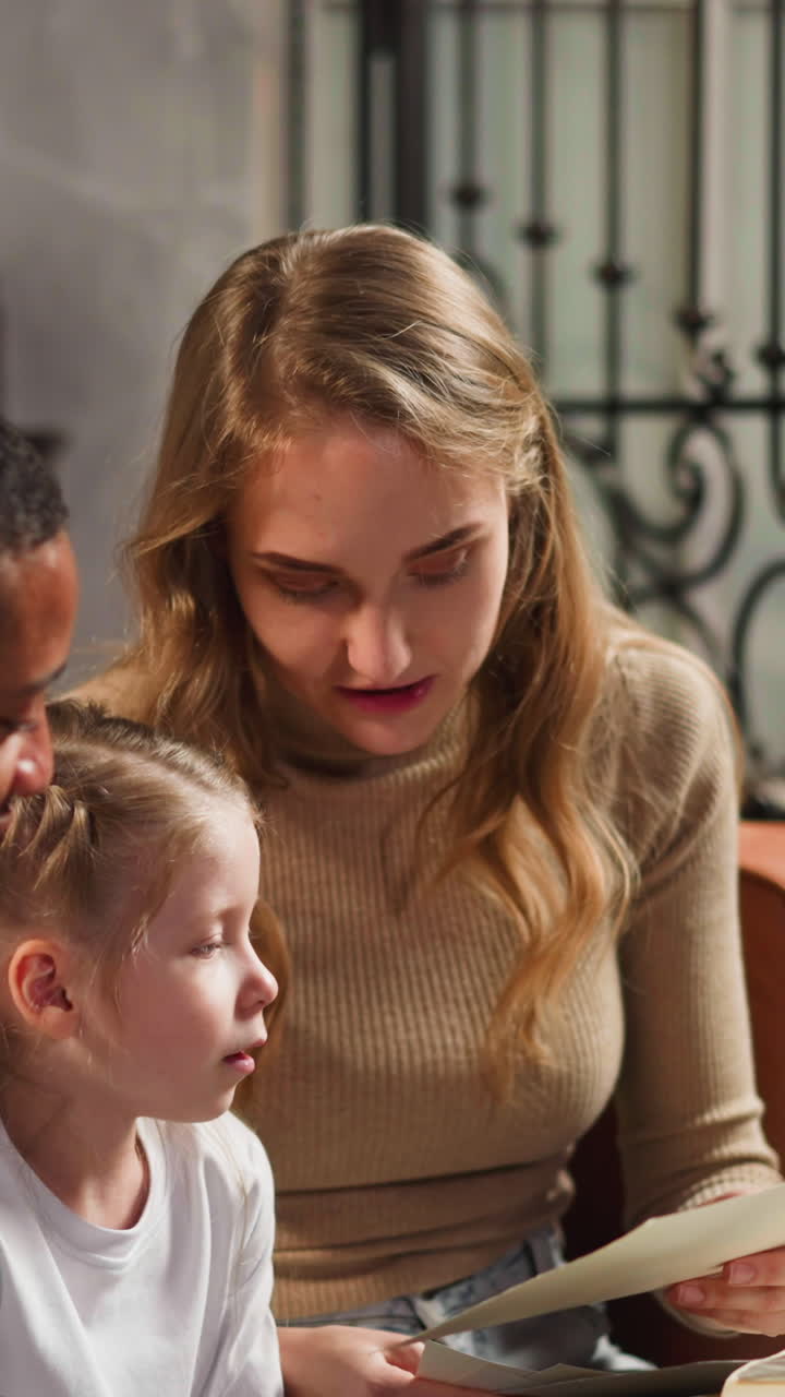 Little girl with plaits asks questions about grandparents to mother holding vintage family pictures with African-American boyfriend at home slow motion