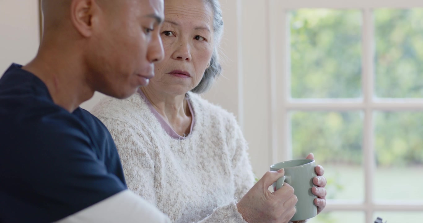 ミントカラーのマグカップを持つ多様な背景を持つシニアの母親と、タブレットとスマートフォンを操作する息子が、大きな窓のそばで写っている